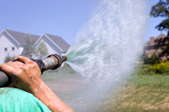 An arm holding a hydrosreed  hose as it sprays materials onto a yard. Two houses and part of a tree in the extreme background.