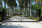 A sliding electric gate in a driveway in front of a large home with palm trees.
