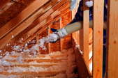 A man spraying blown fiberglass insulation between attic trusses, towards the eaves.