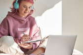 young woman with pink hair and hoodie working on laptop with notebook
