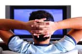 A view from behind a man watching television with his hands behind his head.