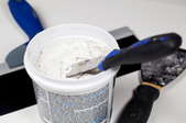 A bucket of spackle with a putty knife in it and another putty knife on the floor next to the bucket