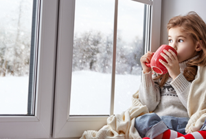 A girl sits by a large window to watch the snowfall
