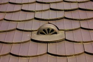 A stylized attic vent surround by matching, brown roofing tiles.