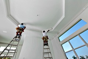 Two men on ladders installing molding around the edges of a tray ceiling.