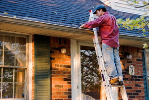 A man standing on a ladder in front of a house cleaning out the gutters. 