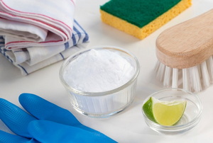 A bunch of cleaning supplies, including a sponge, scrub brush, wash cloths, gloves, baking soda, and a lime wedge.