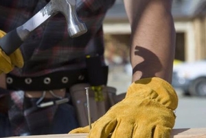 A handyman wearing yellow gloves and hammering nails into a piece of wood outside.