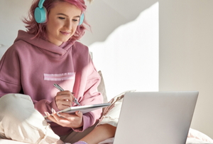 young woman with pink hair and hoodie working on laptop with notebook