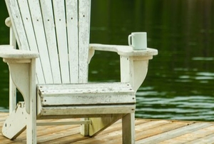 wooden Adirondack chair on a platform by water with a mug