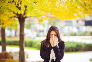 A woman blowing her nose into a tissue with a yellow-leaved tree behind her in the fall.