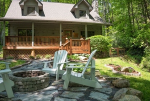 A cabin in the woods with three adirondack chairs around a fire pit.
