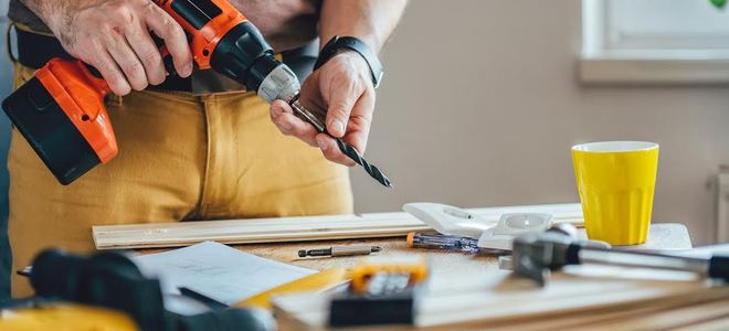 A man holding a cordless drill in front of stacks of wood.