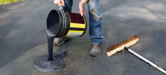 person pouring black sealant onto driveway near brush