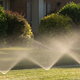 Several bushes line the outside of a house while sprinklers water the yard.