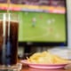 A TV in the background with a football game on, and soda and chips in the foreground. 