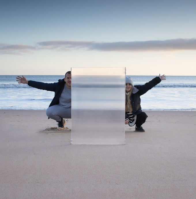 Man and woman pose behind the Invisibility Shield Co's Real Invisibility Shield on a beach.