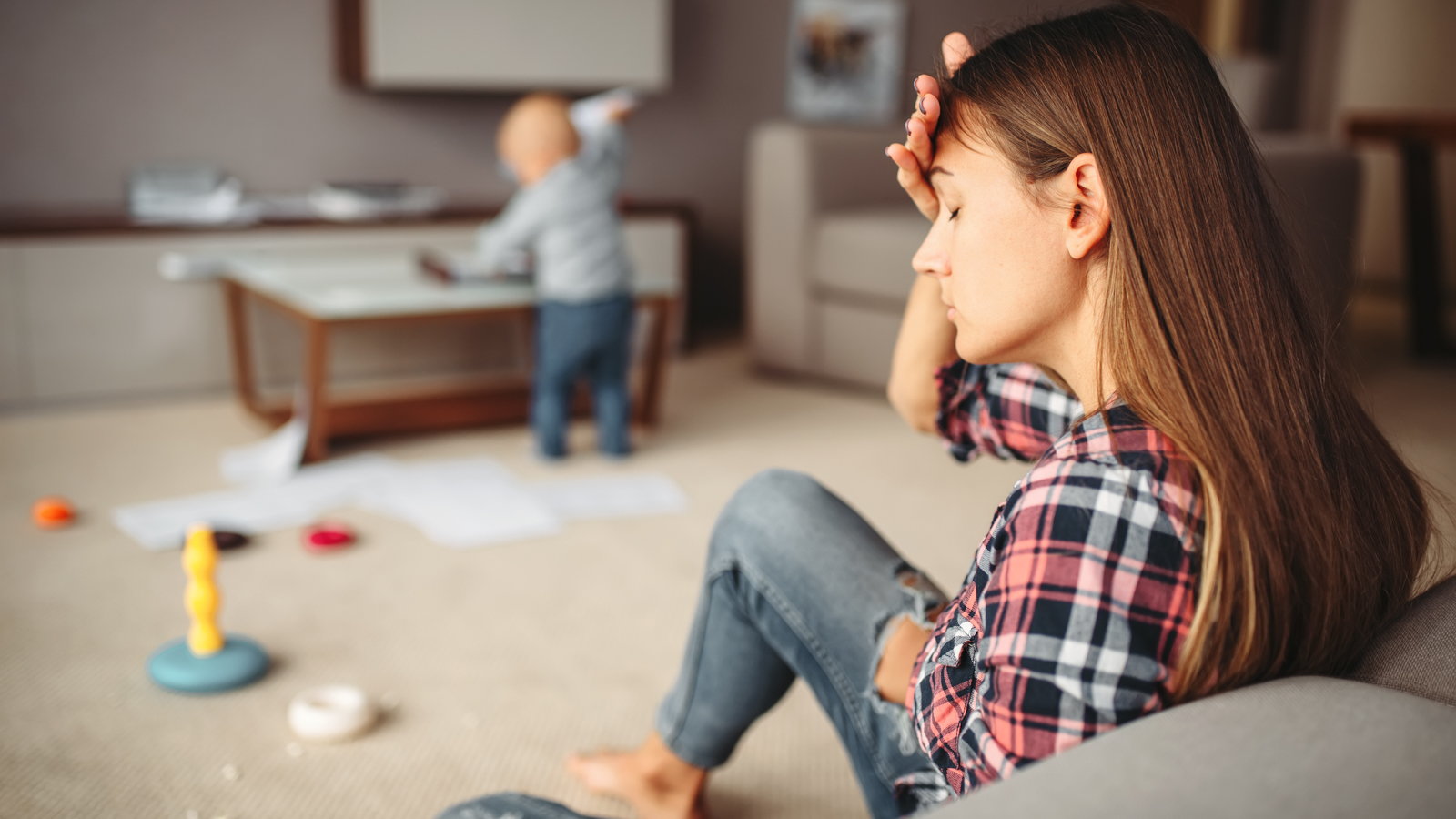 stressed out mom sitting on couch while toddler makes a mess