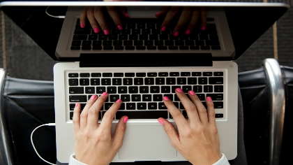 An overhead view of a pair of hands with pink fingernails typing on a keyboard. 