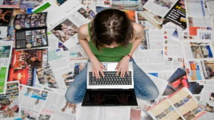An aerial shot of a young woman working on her laptop surrounded by magazines. 